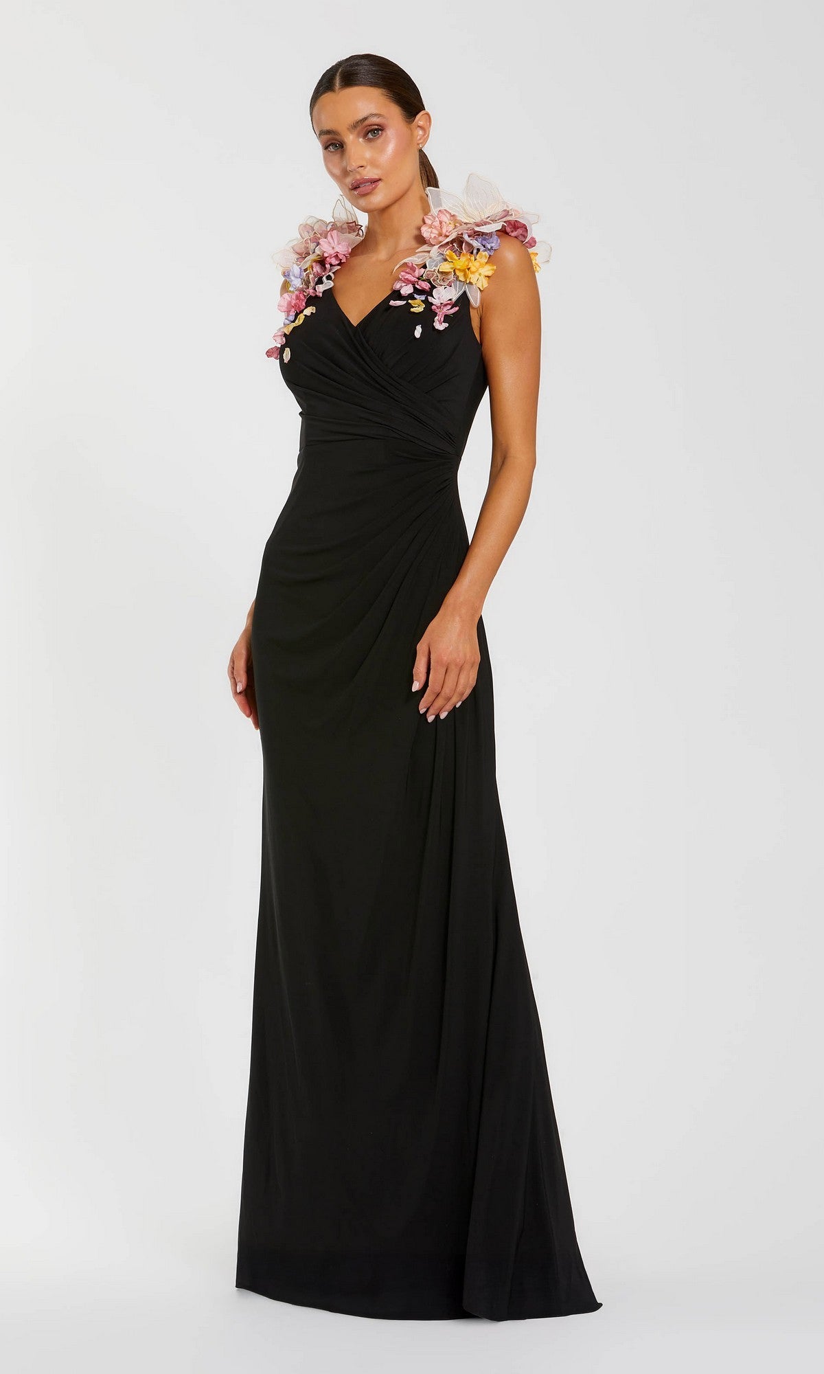 A woman wearing the Mac Duggal 21084 long formal dress in Black Multi, which features a ruched waist and vibrant 3D floral appliques on the shoulders, is posing against a plain white background.
